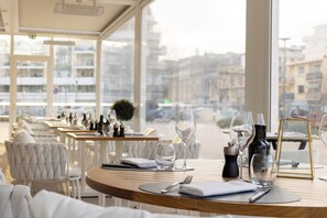 A restaurant with white chairs and tables, some with wine glasses and bottles, and a view of buildings through large windows. - Hotel Indigo Cagnes-sur-Mer by IHG (Cagnes-sur-Mer)