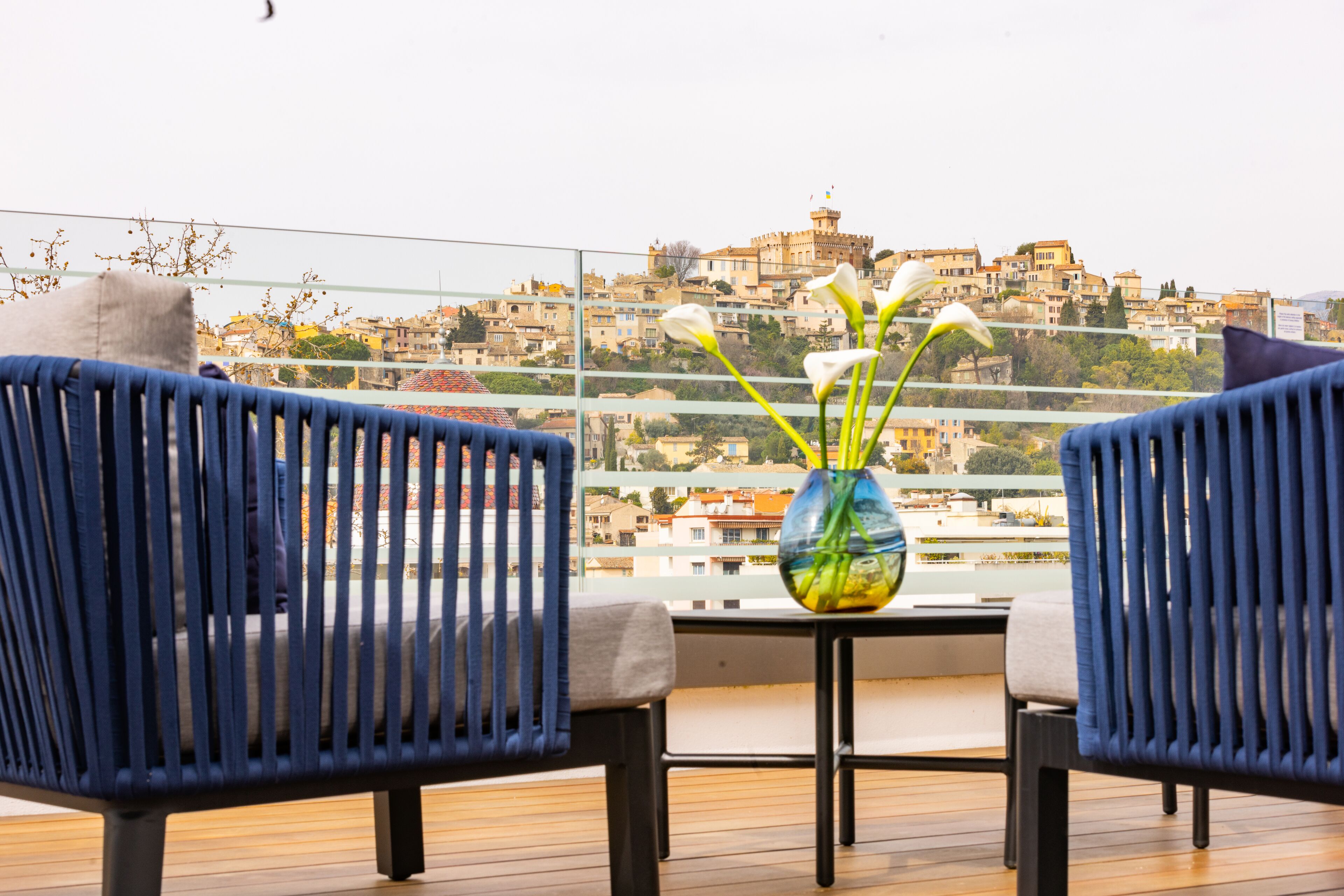 A balcony with two blue chairs and a vase with flowers, overlooking a cityscape with buildings and a castle.