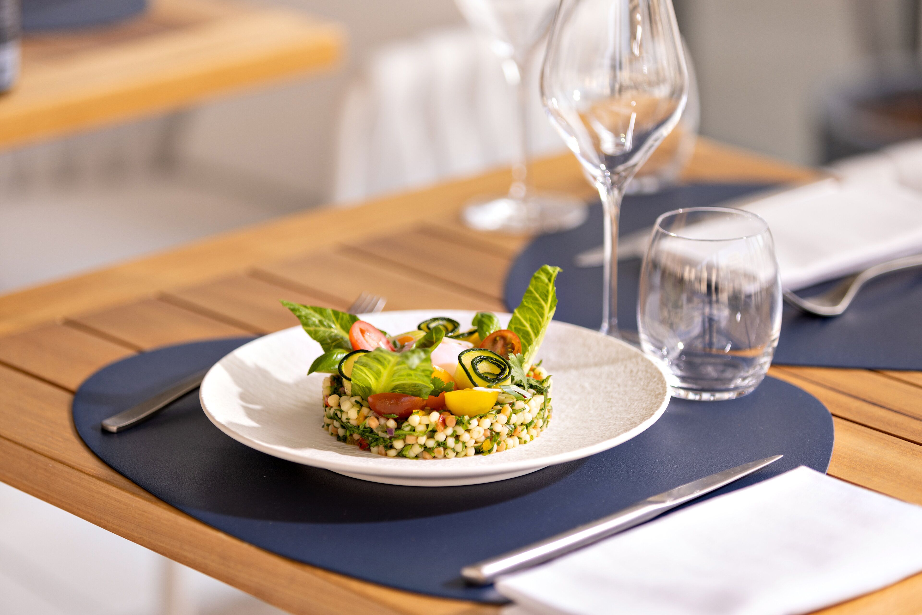 A plated dish with various vegetables and grains, accompanied by a glass of water and a wine glass on a wooden table.