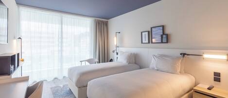 Une chambre d’hôtel avec deux lits, un bureau, une chaise, une télévision et une grande fenêtre avec des rideaux.