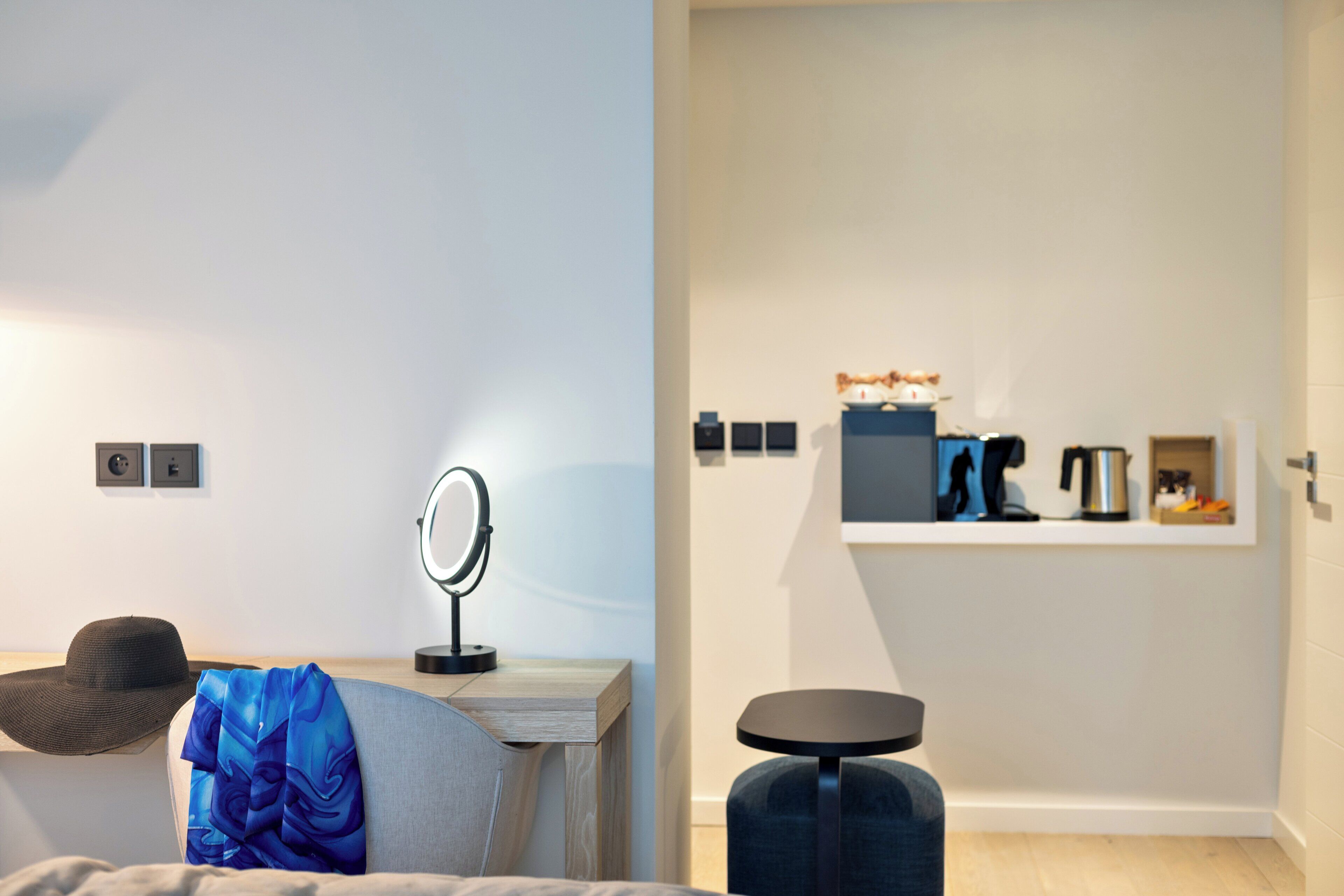 Ein modernes Zimmer mit einem Bett, einem Nachttisch mit Spiegel, einem kleinen Hocker und einem Regal mit verschiedenen Gegenständen.