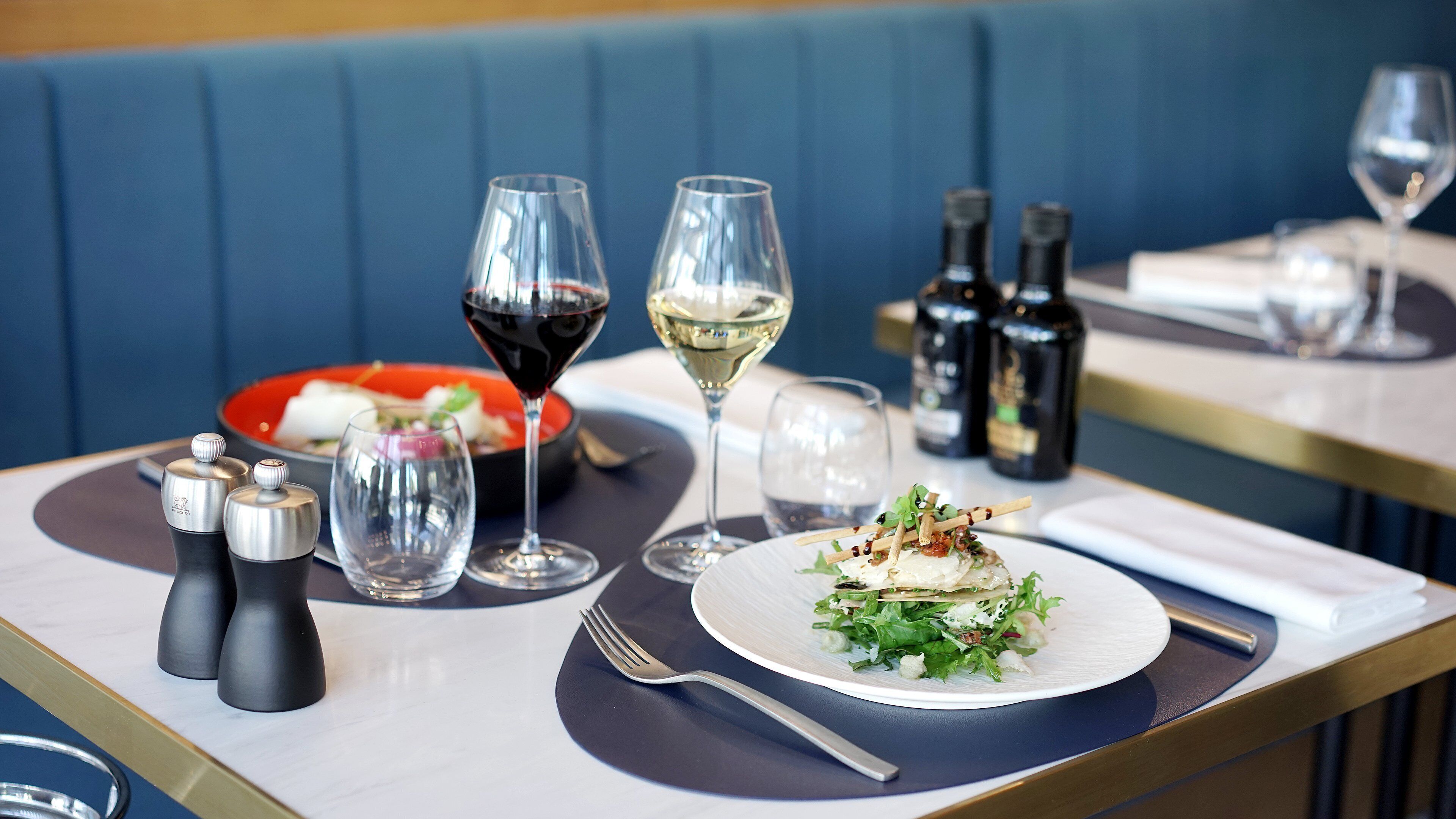 Une table à manger avec une assiette de nourriture, deux verres à vin, des salières et des poivrières, et deux bouteilles de vin.