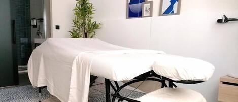 Une salle d’examen médical équipée d’un lit, d’un petit tabouret, d’une plante en pot et d’un miroir fixé au mur.