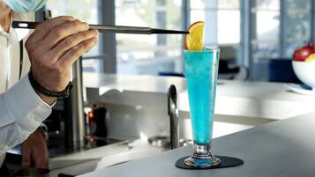 Una persona sosteniendo un cuchillo sobre un vaso alto con un líquido azul y una rodaja de naranja como adorno.
