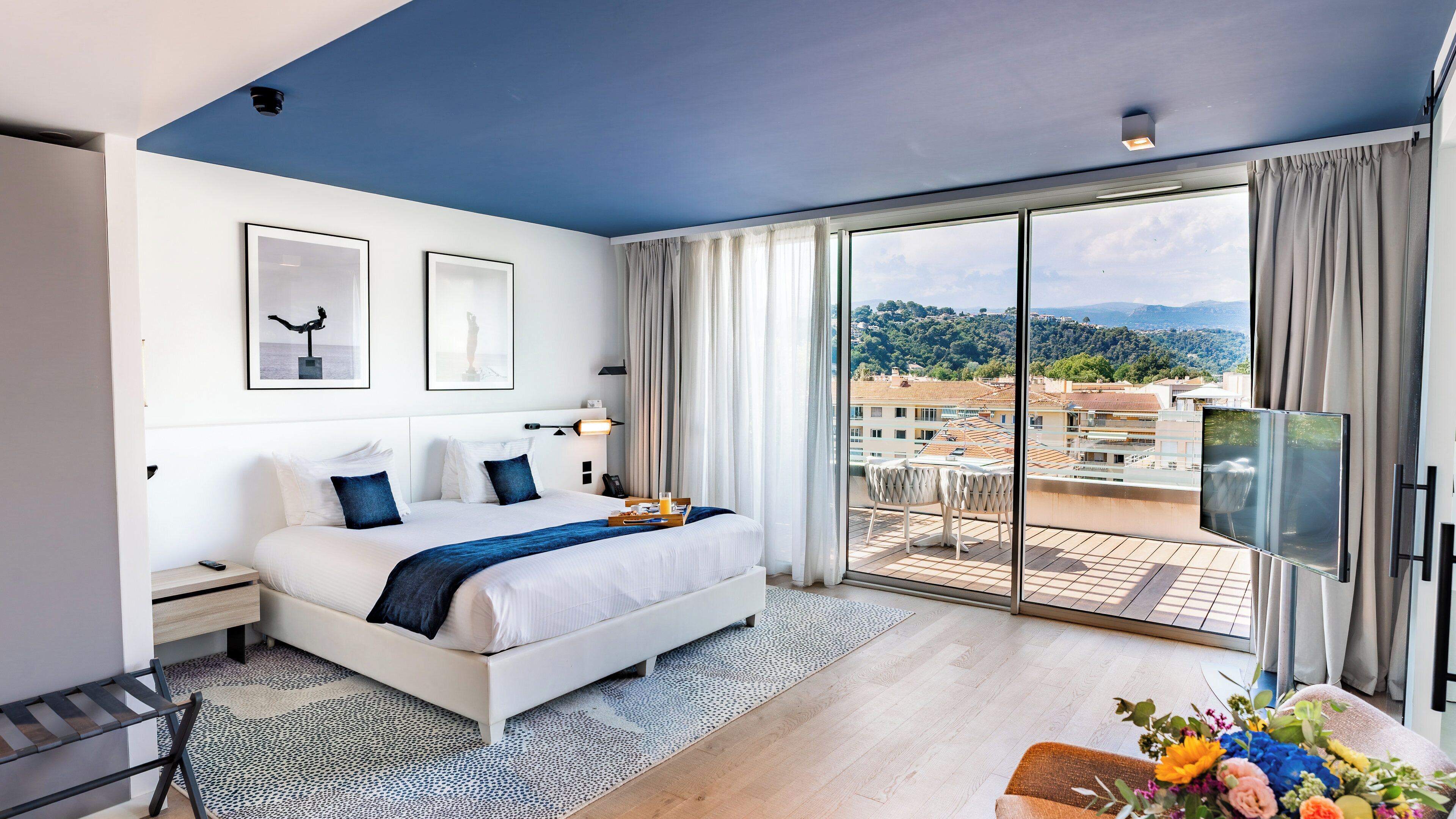 Une chambre d’hôtel moderne dotée d’un grand lit, d’une télévision à écran plat, d’un balcon offrant une vue sur la ville et d’œuvres d’art accrochées aux murs.