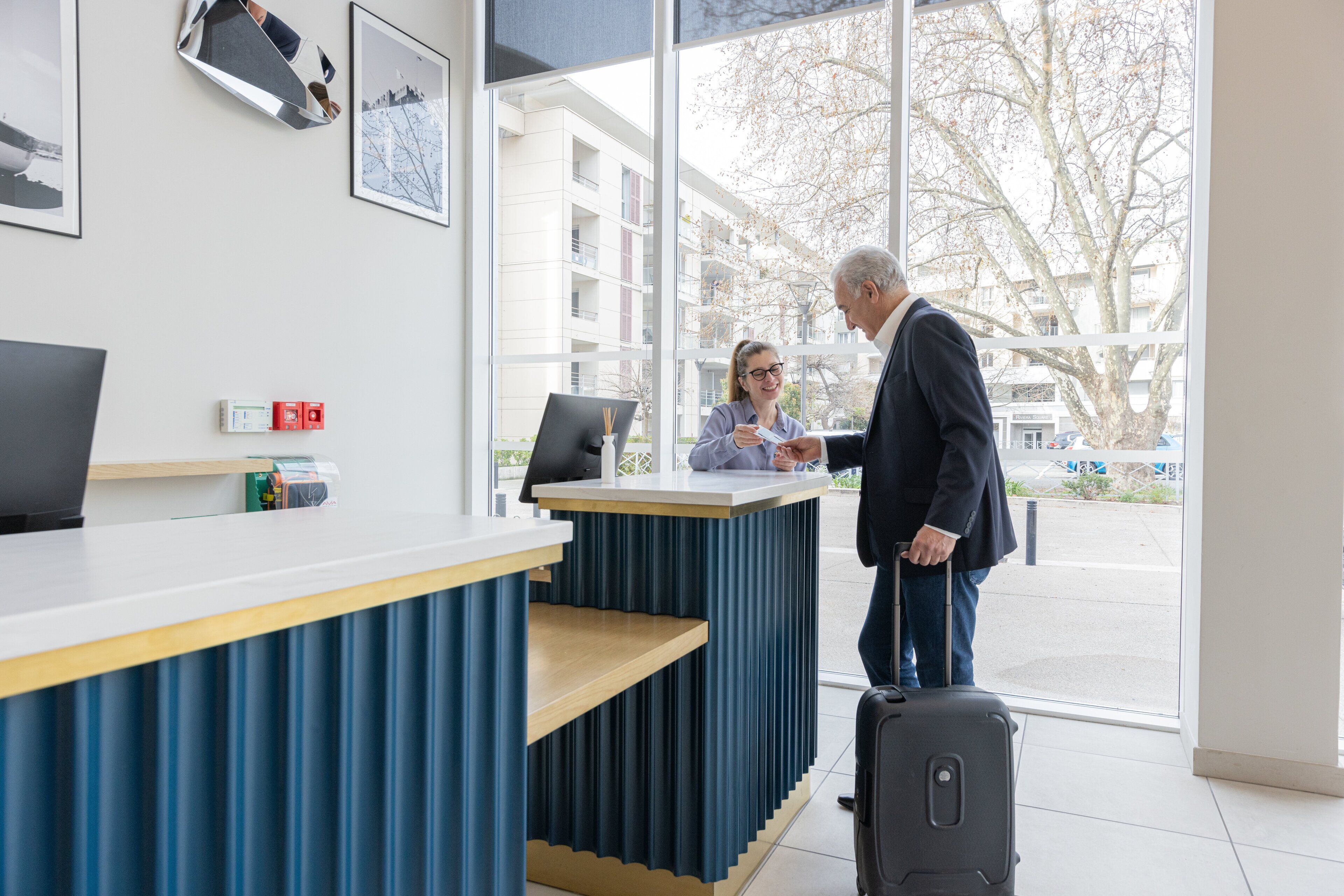 Un comptoir d’accueil avec un ordinateur, un mur d’accent bleu et une fenêtre donnant sur des bâtiments et des arbres.