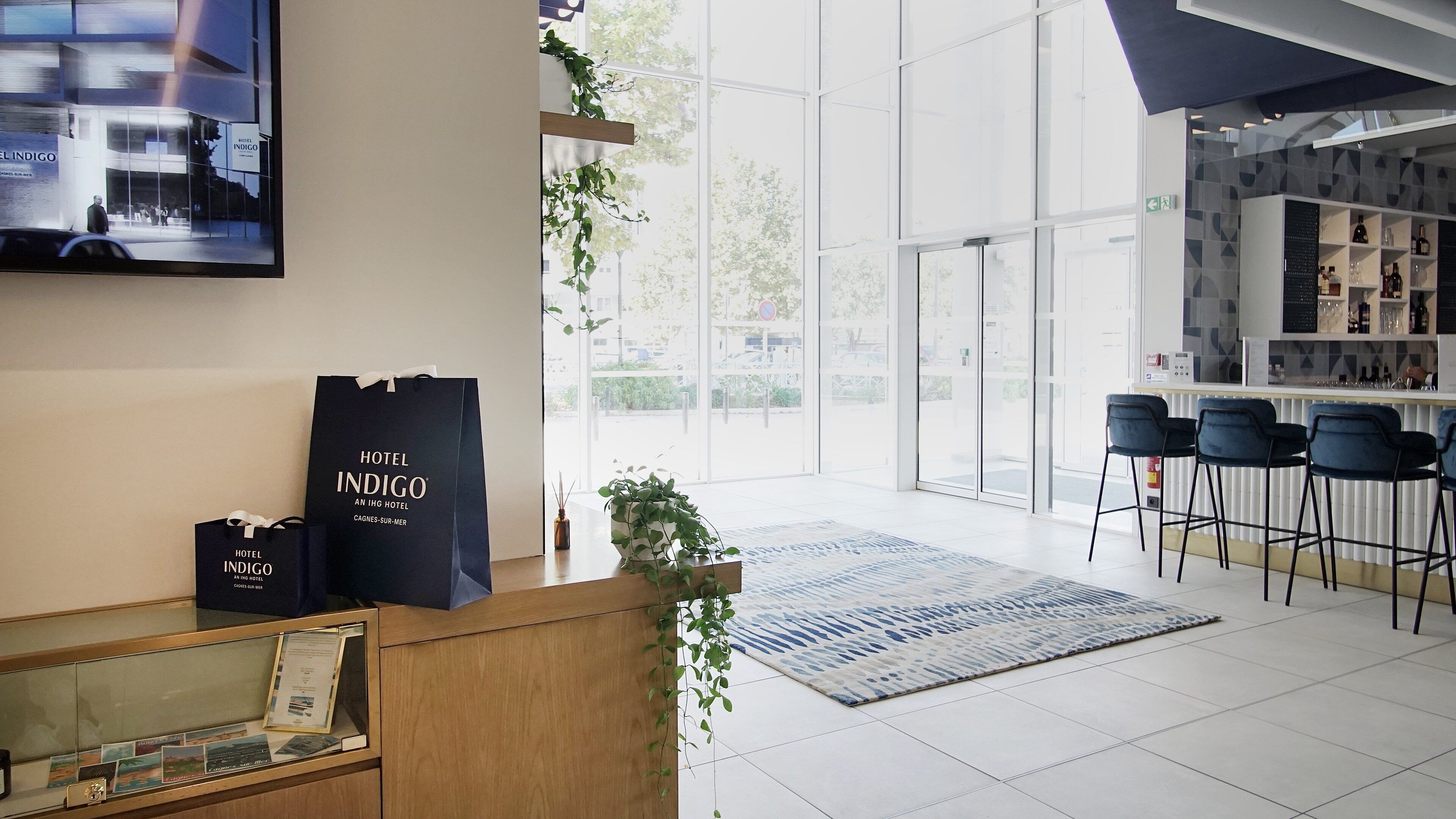 A hotel reception area with a modern design, featuring a reception desk, a bar area with stools, and a large window providing a view of the outdoors.