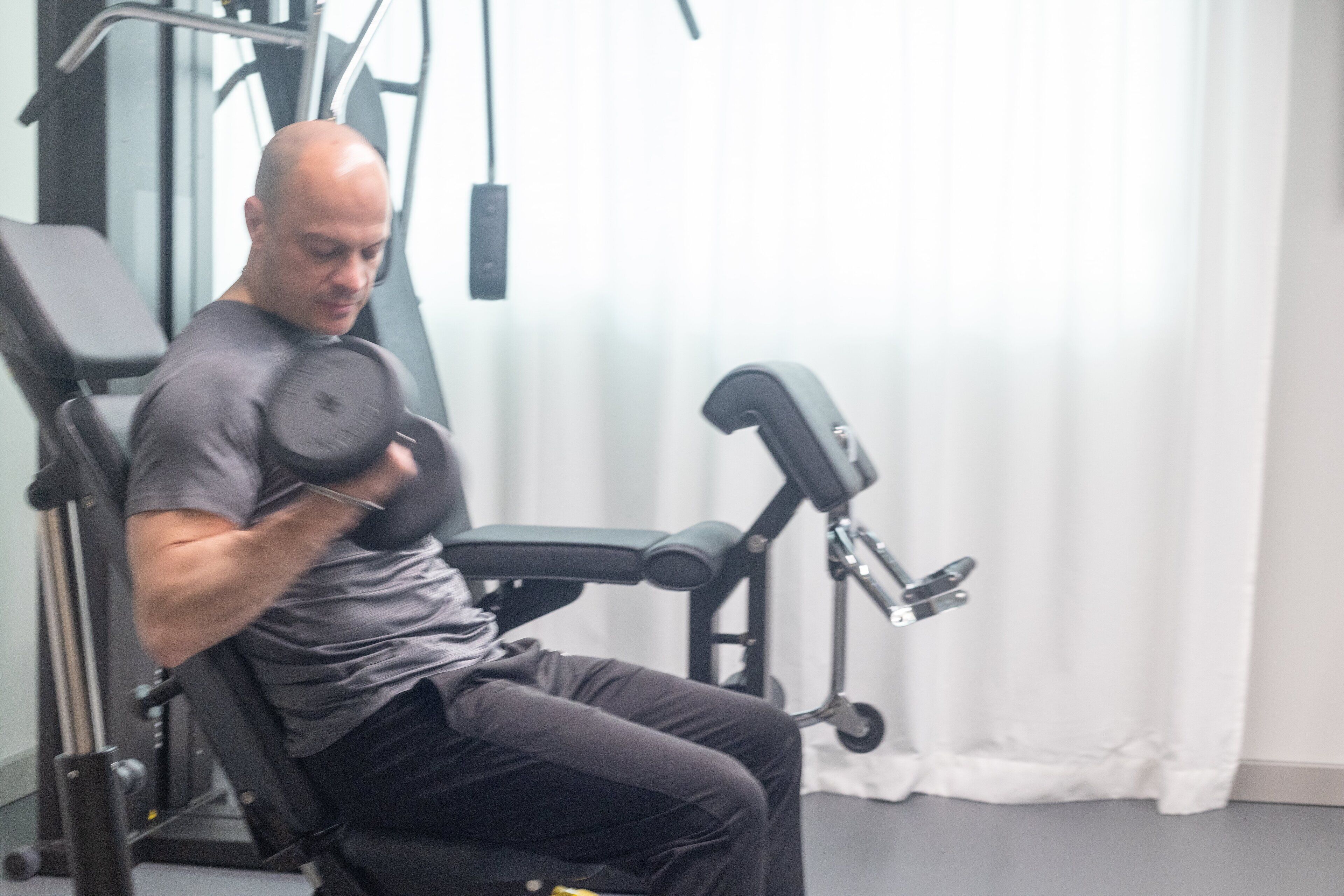 Un hombre usando una máquina de pesas sentada en un gimnasio con equipo de ejercicio y una cortina blanca al fondo.