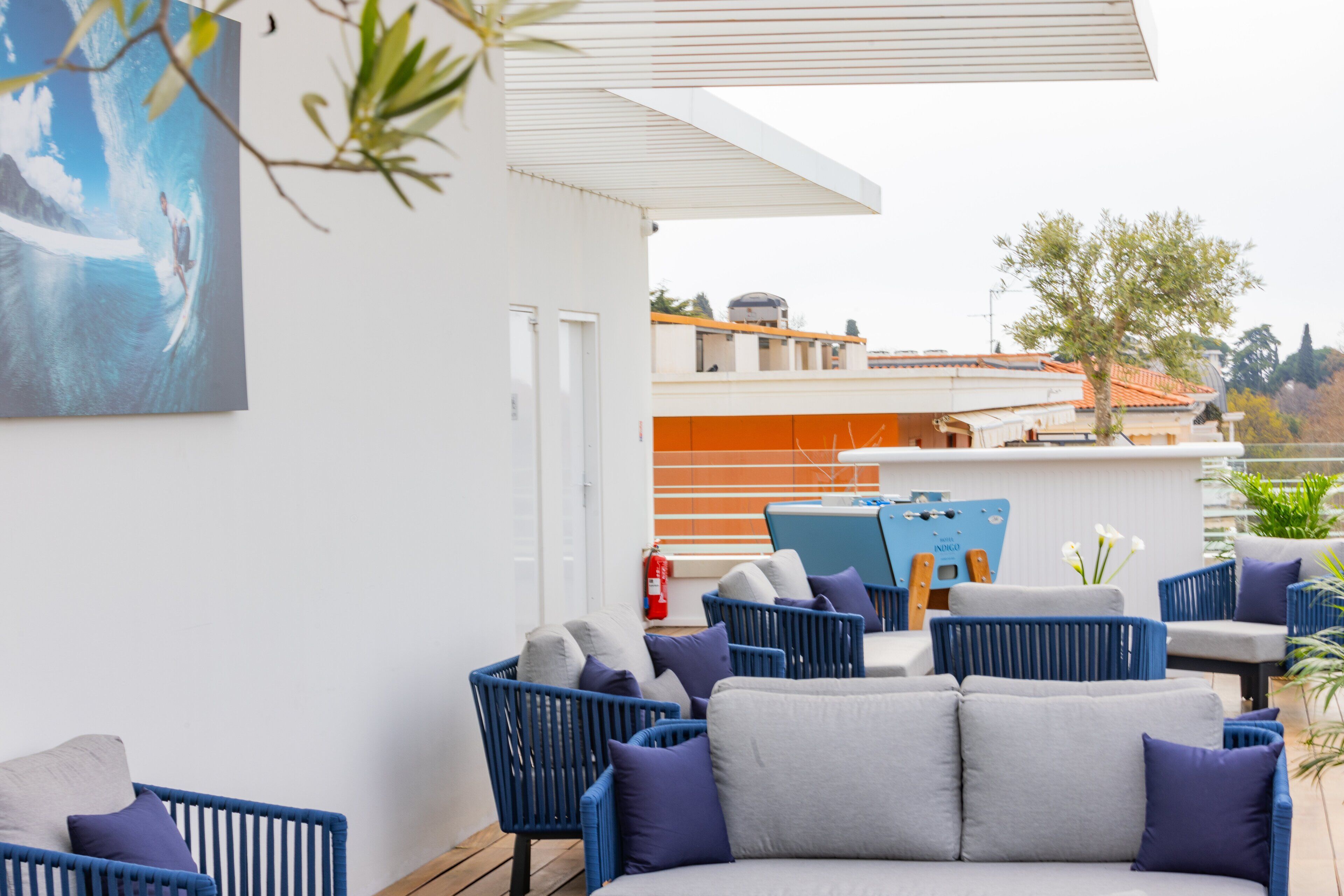 Un espace extérieur moderne aménagé avec des meubles bleus et gris, un mur blanc orné d’un tableau, et une vue sur un bâtiment en arrière-plan.