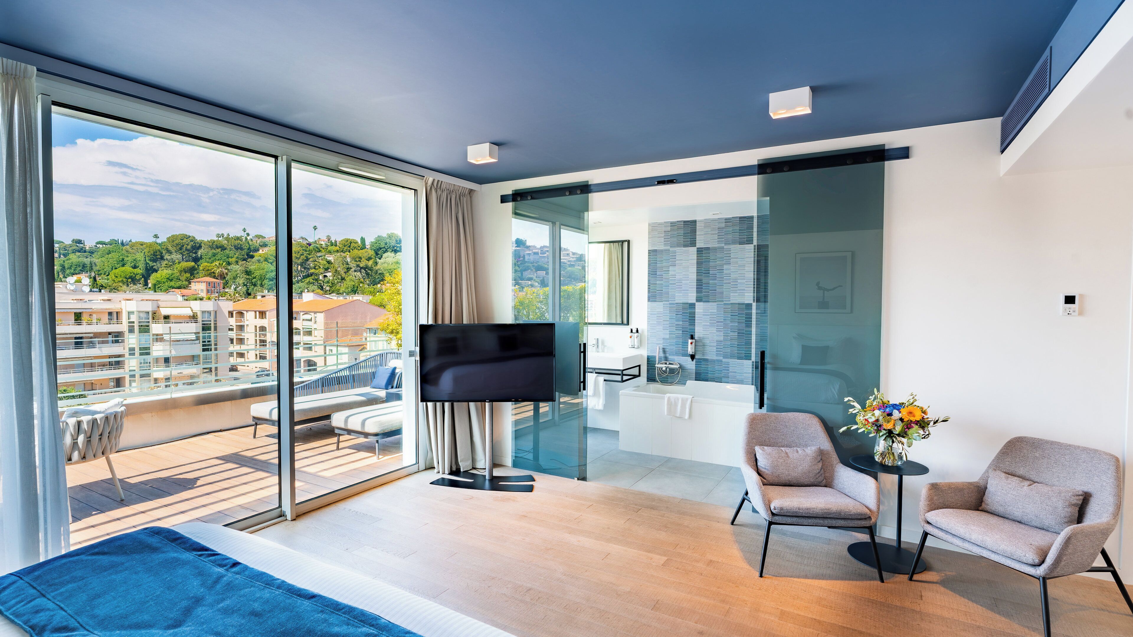 Habitación de hotel moderna con un gran ventanal corredero al balcón, un televisor de pantalla plana, un sofá, dos sillones y una mesita con un jarrón de flores.