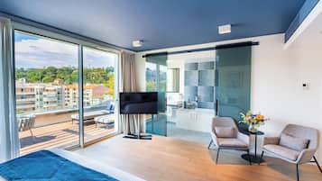 Habitación de hotel moderna con un gran ventanal corredero al balcón, un televisor de pantalla plana, un sofá, dos sillones y una mesita con un jarrón de flores.