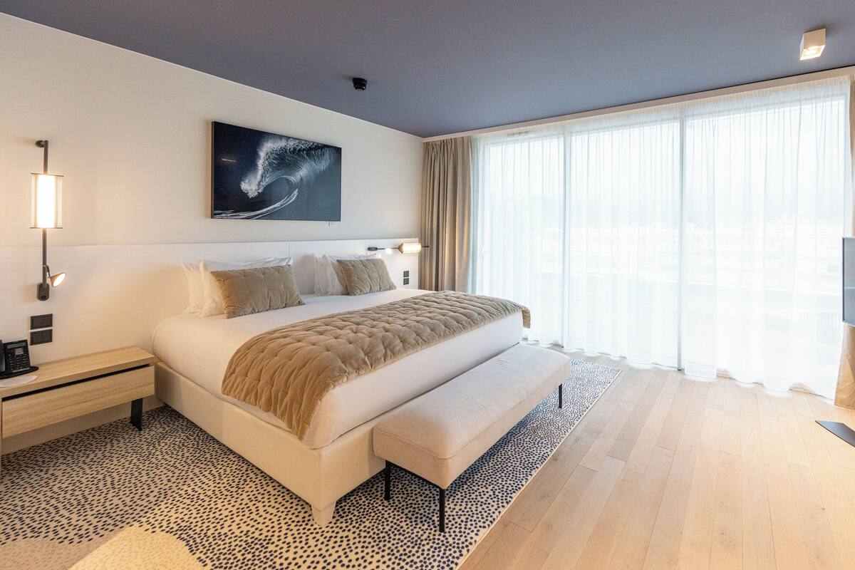 A modern hotel room with a large bed, a flat-screen TV, a bedside table with a lamp, and a large window with sheer curtains.