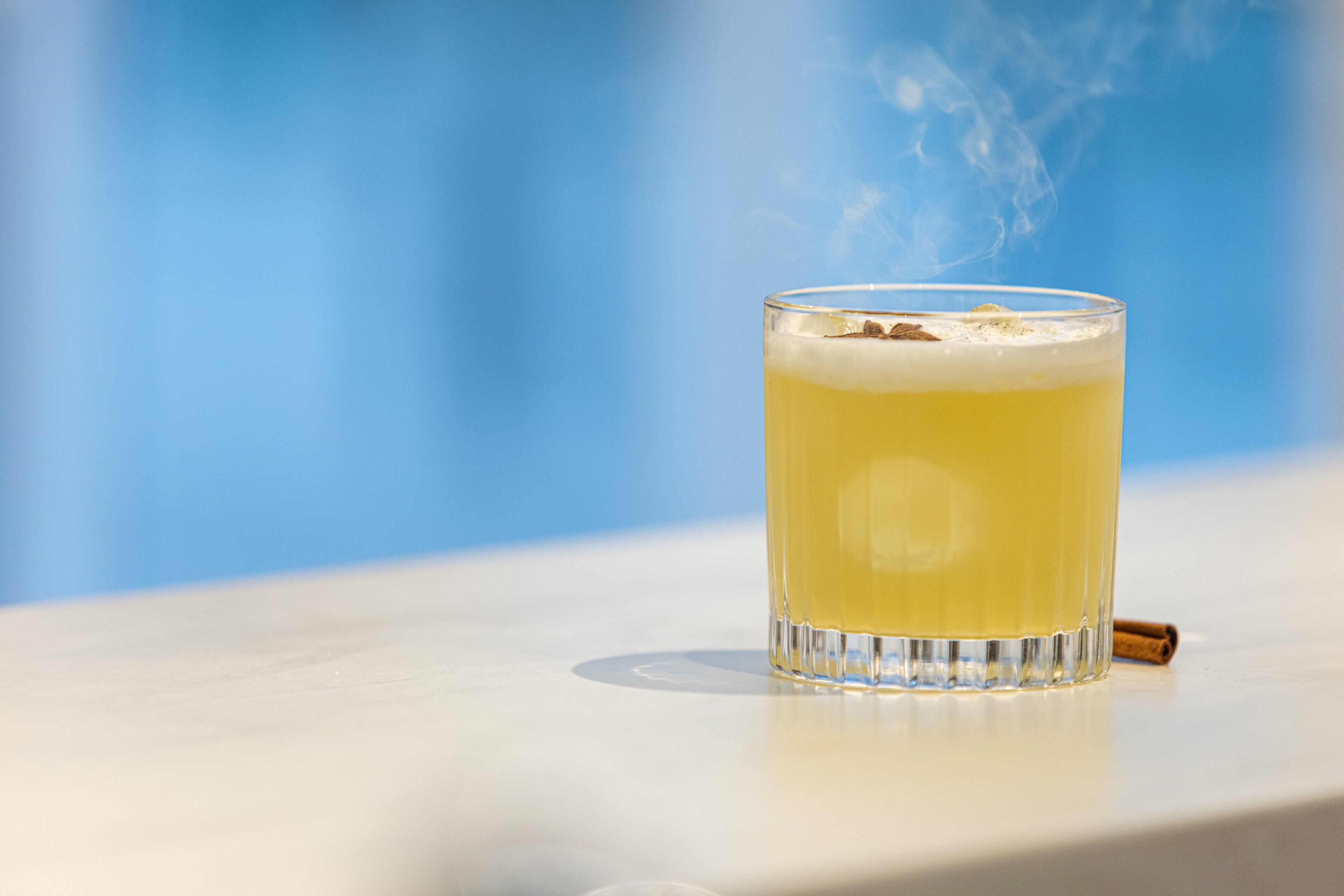 A glass of yellow beverage with a cinnamon stick on a white surface.