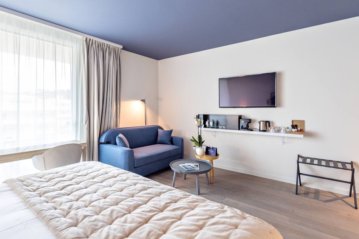 A modern hotel room with a large bed, a blue sofa, a small round table with a lamp, a TV mounted on the wall, and a shelf with various items.