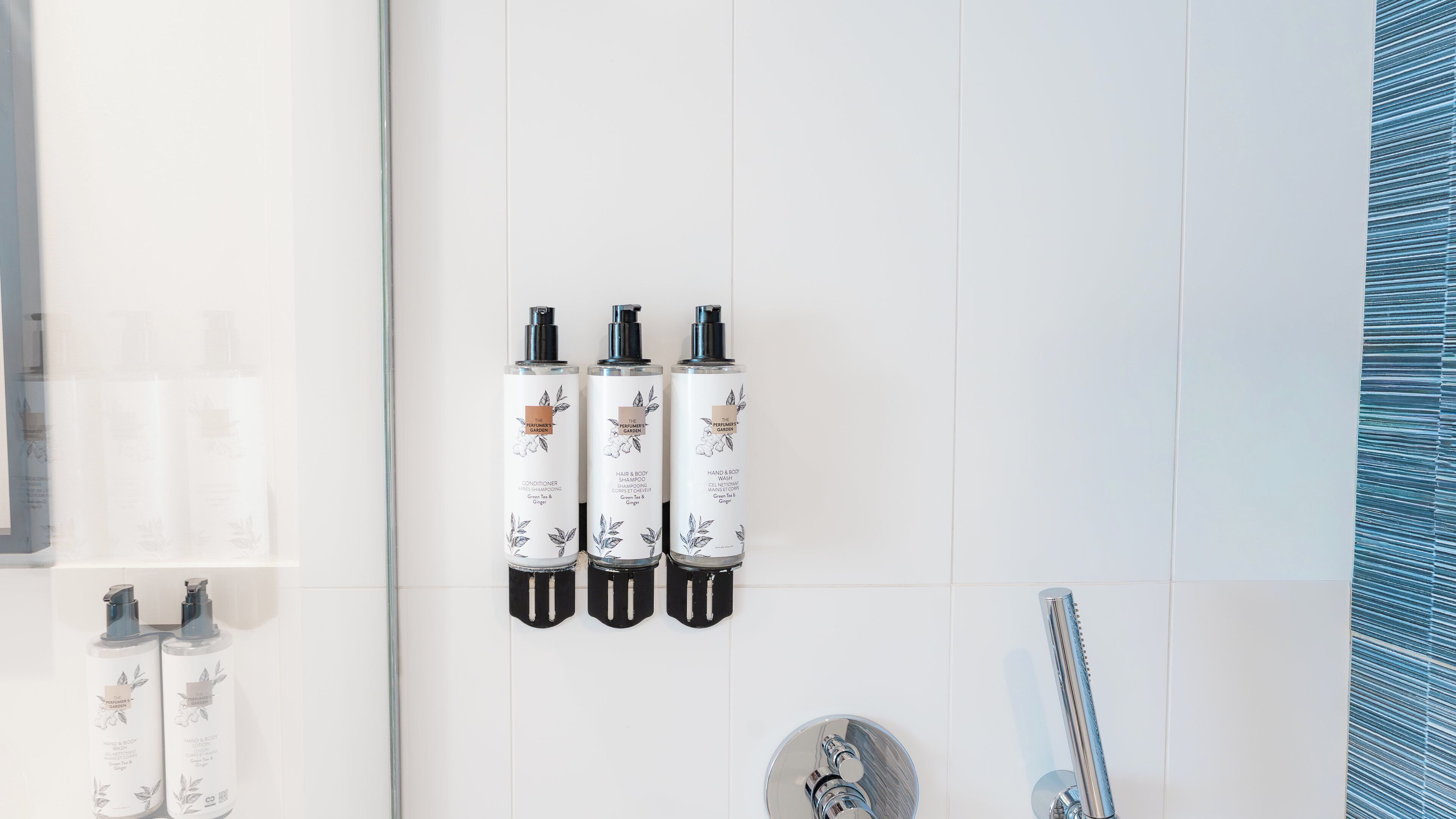 Een badkamer met witte tegels, een douche en drie flessen hand- en douchezeep aan de muur.
