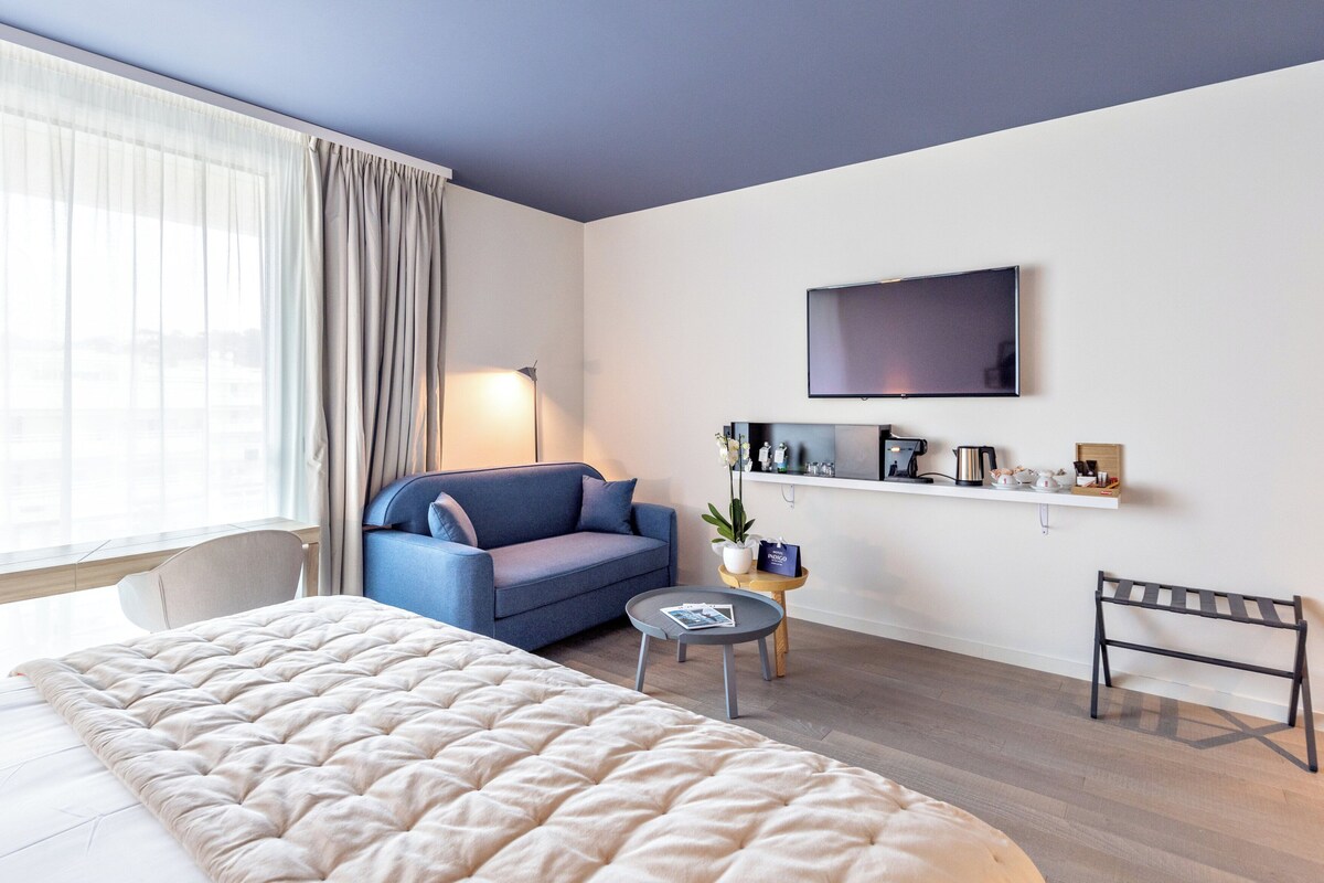 A modern hotel room with a large bed, a blue sofa, a small round table with a lamp, a TV mounted on the wall, and a shelf with various items.