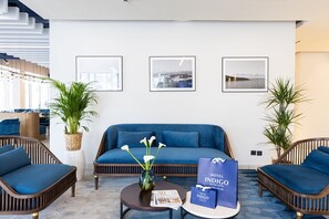 A modern hotel lobby with a blue sofa, wicker chairs, a coffee table with a vase and magazine, and framed pictures on the wall. - Hotel Indigo Cagnes-sur-Mer by IHG (Cagnes-sur-Mer)
