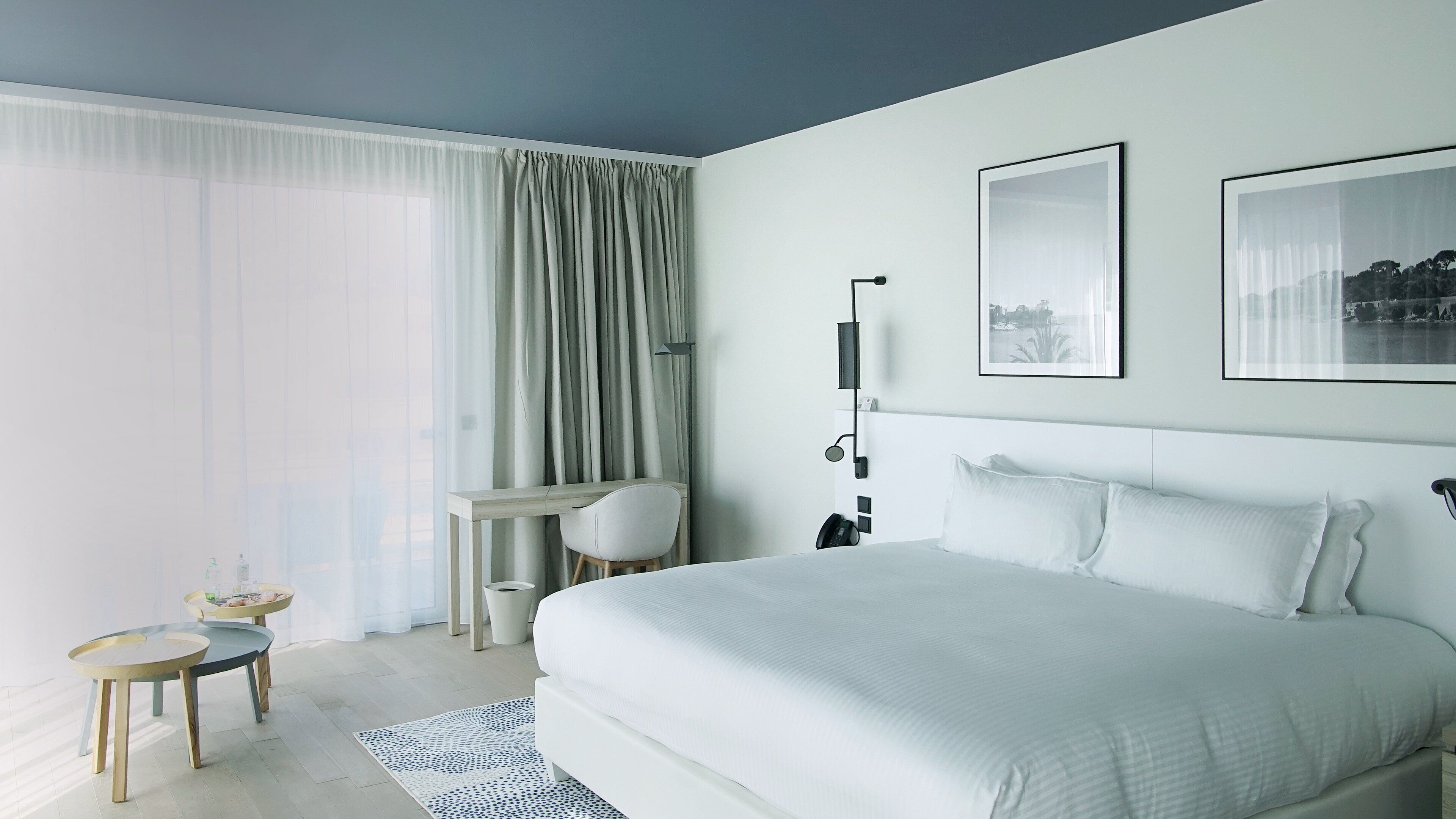 A modern hotel room with a large bed, a desk with a chair, a small table with a lamp, and a large window with curtains.