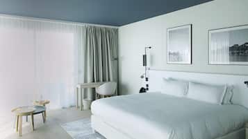 A modern hotel room with a large bed, a desk with a chair, a small table with a lamp, and a large window with curtains.