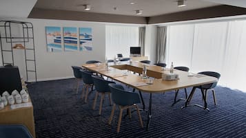 Et mødelokale med et langt bord, stole, en projektor og et skærm. Der er vinduer med gardiner, et klædestativ og en væg med kunst.