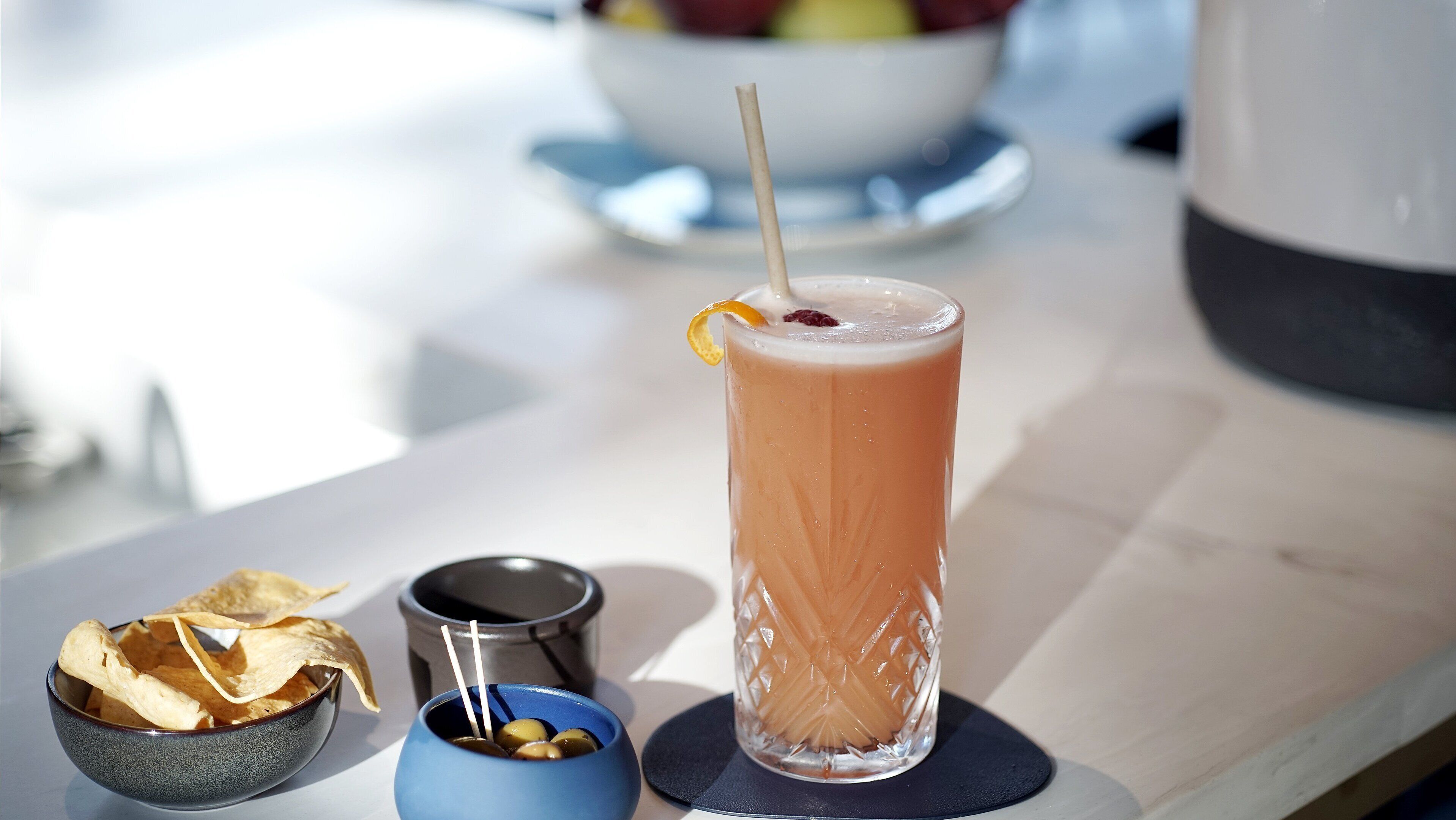 A tall glass with a straw, a bowl of chips, a small bowl of olives, and a coffee pot on a table.