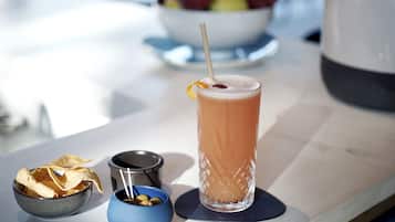 Un vaso alto con pajita, un cuenco de papas fritas, un bol pequeño de aceitunas y una cafetera sobre una mesa.