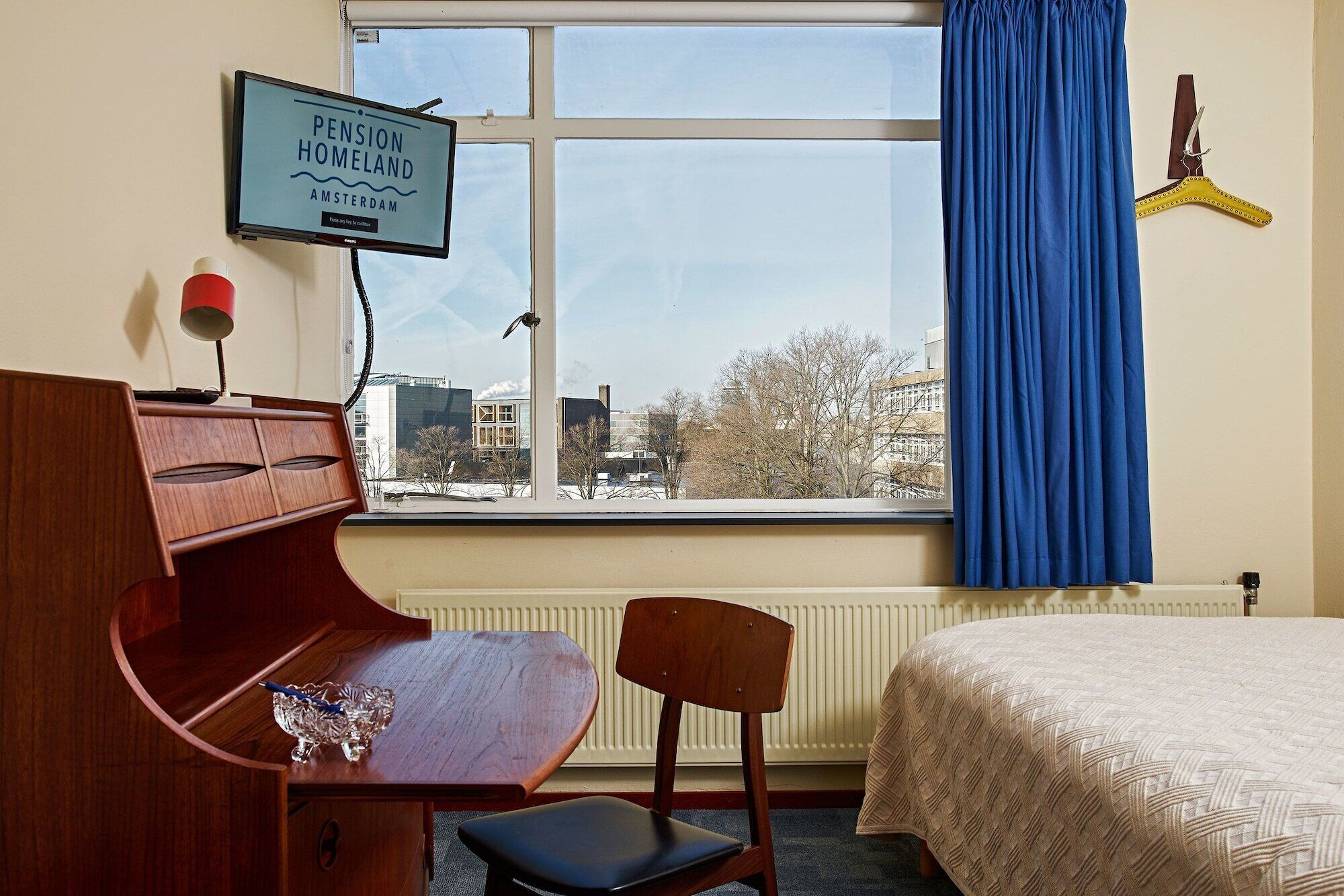 Een kamer met een houten bureau, een stoel, een bed, een raam met uitzicht op gebouwen, en een tv waarop 'Pension Homeland Amsterdam' staat.