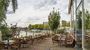 An outdoor patio with wicker furniture and tables, overlooking a river with boats and buildings in the background. - Pension Homeland (Amsterdam)