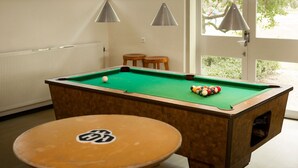 A pool table with balls, a pool cue, and a table with a logo. There are two hanging lamps and a window with a view of greenery. - Pension Homeland (Amsterdam)
