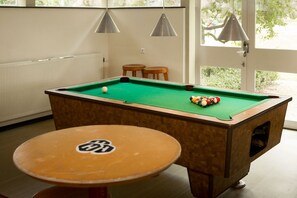 A pool table with balls, a pool cue, and a table with a logo. There are two hanging lamps and a window with a view of greenery. - Pension Homeland (Amsterdam)