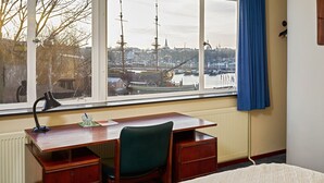 A room with a wooden desk, a chair, a bed, and a window with a view of a harbor and ships. - Pension Homeland (Amsterdam)