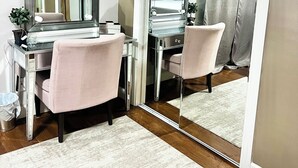 A room with a vanity, mirror, and two chairs, a desk with a computer, and a large mirror reflecting the room.