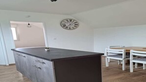 Une cuisine moderne avec un plan de travail foncé et des armoires en bois, un coin repas avec des chaises blanches et une table en bois, et une horloge murale.