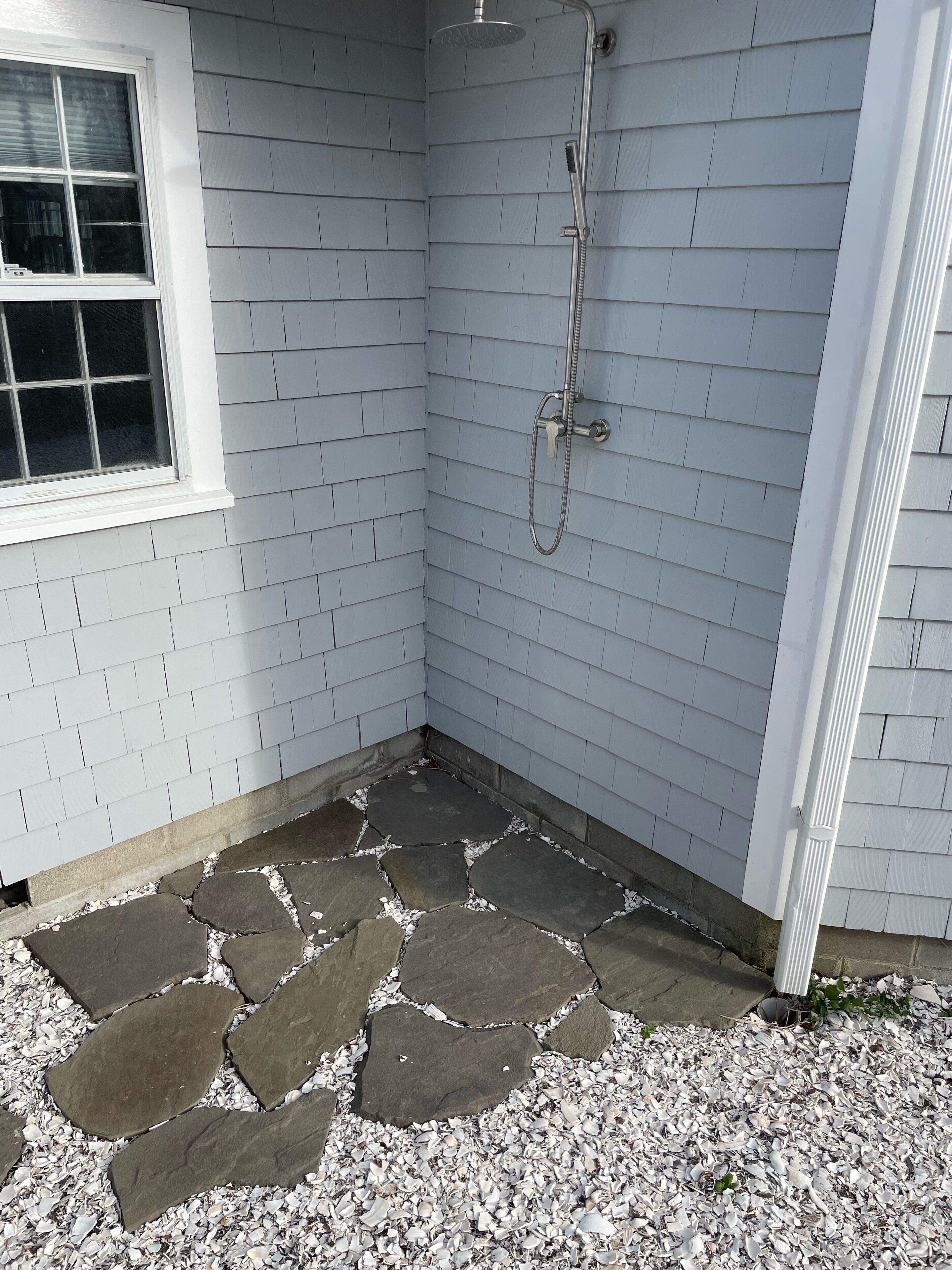 A corner of a room with white siding, a window, and a shower area with a hose.