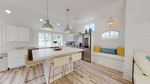 Une cuisine moderne avec des meubles blancs, un îlot central doté de tabourets de bar, un réfrigérateur en acier inoxydable et des luminaires suspendus.
