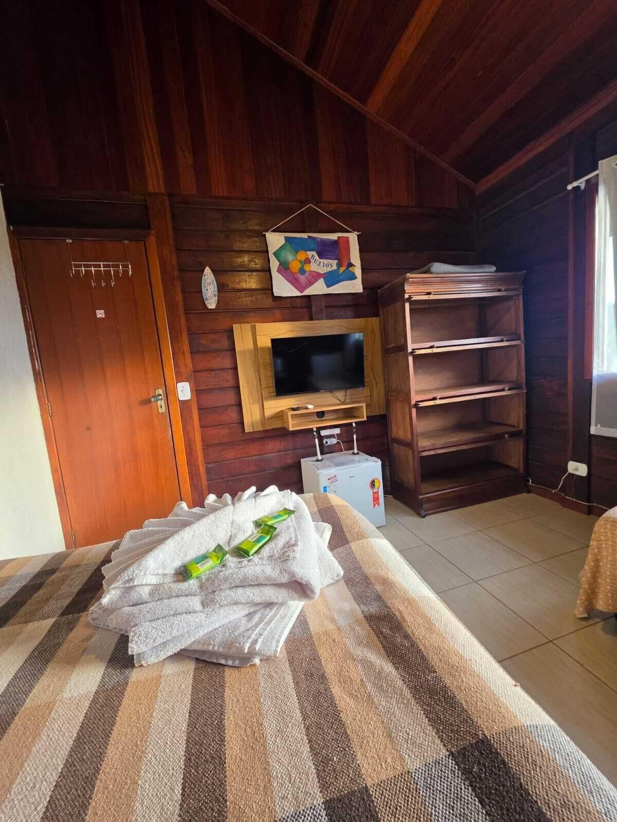 A room with a wooden ceiling, a television mounted on a wooden frame, a door with a towel rack, and a bed with a striped bedspread.