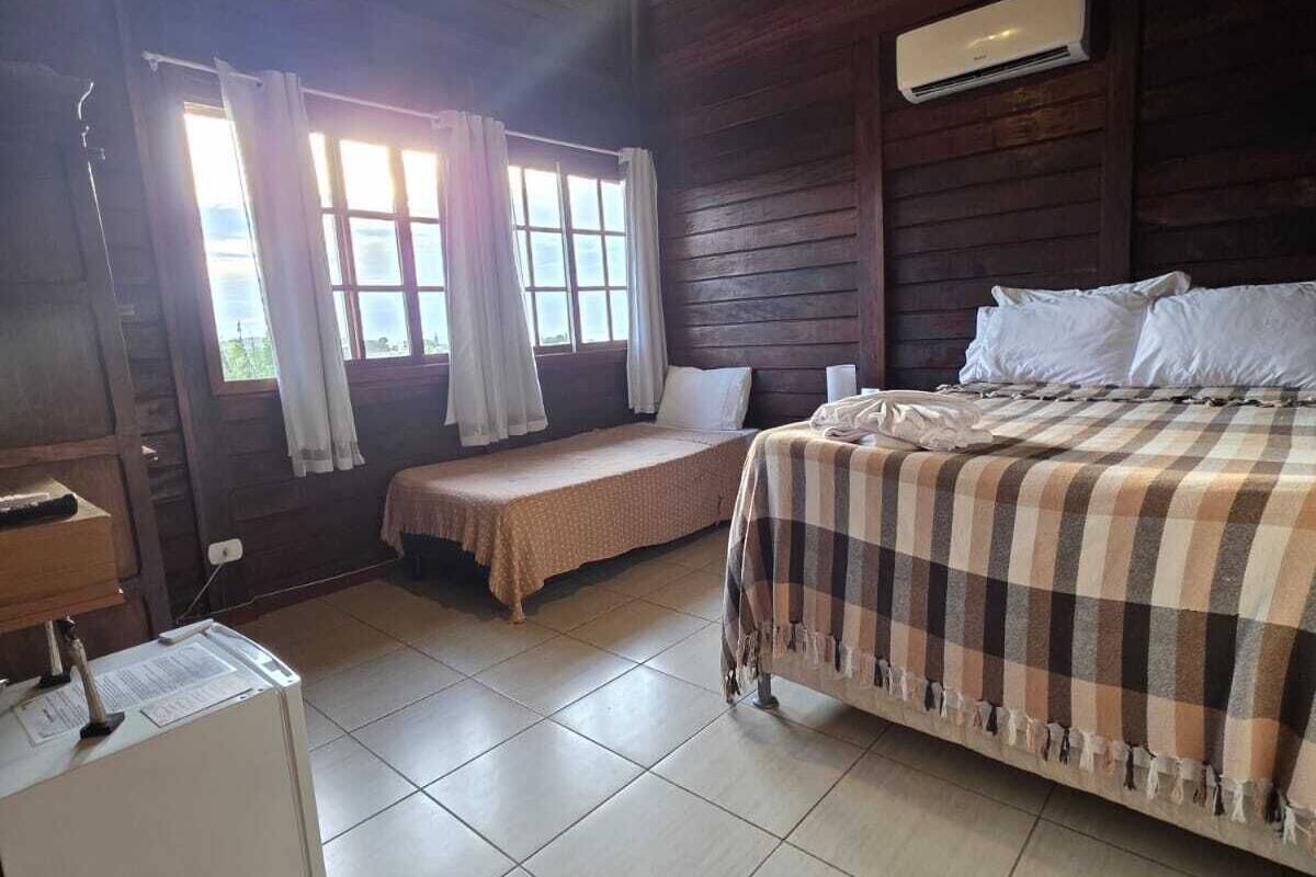 Ein Zimmer mit einem Bett, einem kleinen Tisch, einem Schreibtisch, einem Fenster mit Vorhängen und einer Deckenleuchte.