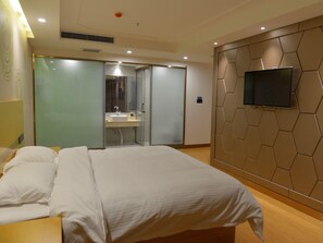 Habitación de hotel con cama, televisor instalado en una pared hexagonal y baño visible a través de una puerta de cristal.