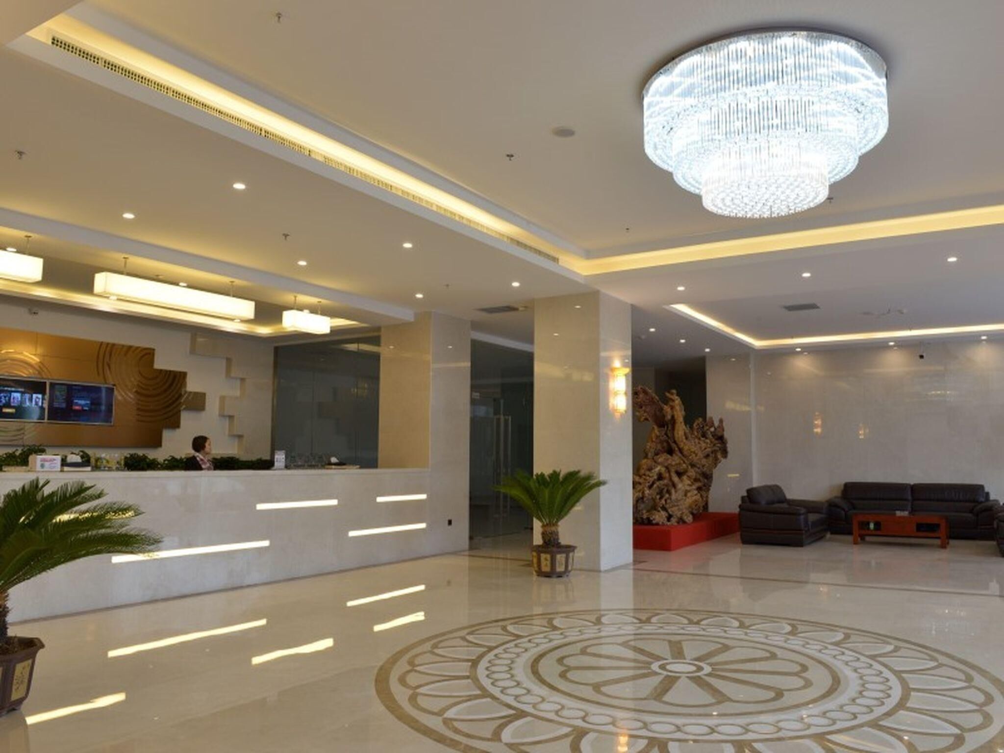 A modern hotel lobby with a reception desk, a chandelier, a patterned floor design, and a seating area with sofas.