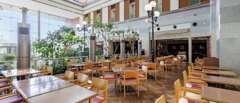A spacious indoor dining area with multiple tables and chairs, a high ceiling with a glass roof, and a view of the outside through large windows.