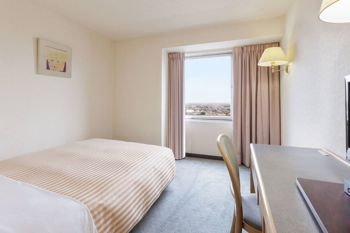 Une chambre d’hôtel avec un lit, un bureau, une chaise, une fenêtre avec des rideaux et une lampe.