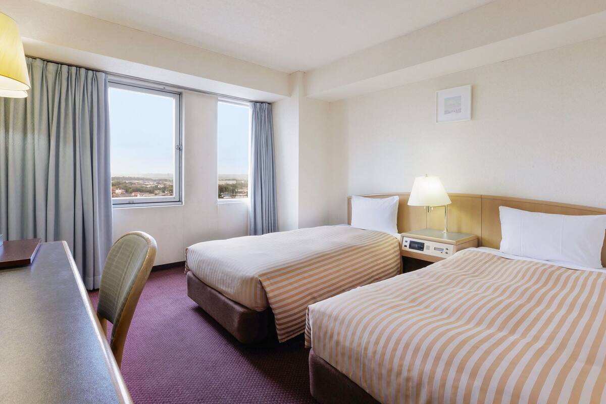 Une chambre d’hôtel avec deux lits, un bureau, une chaise, une lampe et une vue sur la ville.