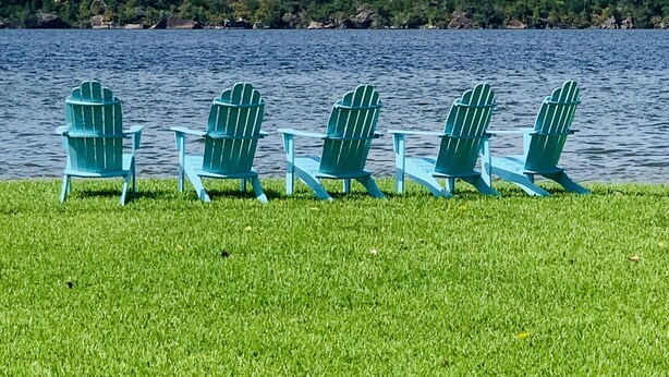 Een rij groene Adirondack-stoelen met uitzicht op een meer, onder een heldere lucht en met een verre oever.