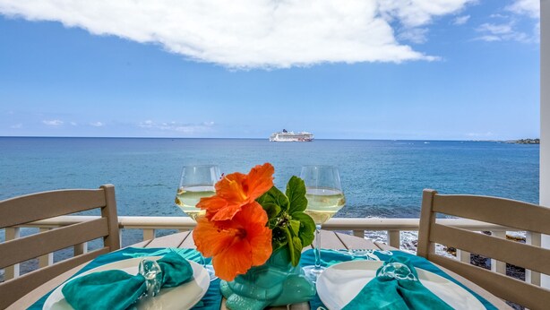 Une table ornĂ©e dâun bouquet de fleurs, de deux verres de vin blanc et dâune nappe aux motifs turquoise et blancs. On aperçoit un navire de croisiĂšre au loin, de lâautre cĂŽtĂ© de lâeau.