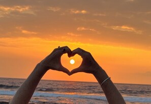 Hands forming a heart shape around the sun at sunset over the ocean.