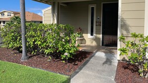 A two-story house with a covered porch, a small garden with shrubs and a potted plant, and a concrete pathway leading to the entrance. - Stunning ocean view Ali'i Cove J-1 (Kailua Kona)