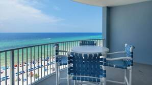 A balcony with a round table and two chairs, overlooking a beach with umbrellas and people. - Twin Palms 1103 (Panama City Beach)
