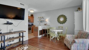 A living room with a television, wooden furniture, a dining table with chairs, and a kitchen in the background. - Twin Palms 1103 (Panama City Beach)