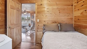 Habitación con paneles de madera, una cama, una silla blanca y una puerta que da a otra habitación.