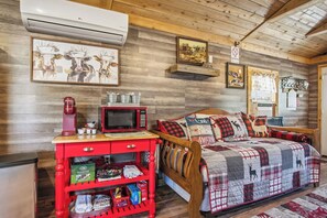 A cozy room with a wooden ceiling, a bed with a quilted blanket, a red kitchen counter with a microwave, and a red cabinet with various items. - RRR Cabin on the Medina | 1800 Feet River | Deer (Medina)